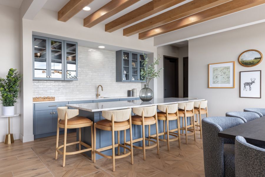 Modern kitchen with blue cabinets, wood beams, bar stools, a vase on the counter, and framed art on a light-colored wall.