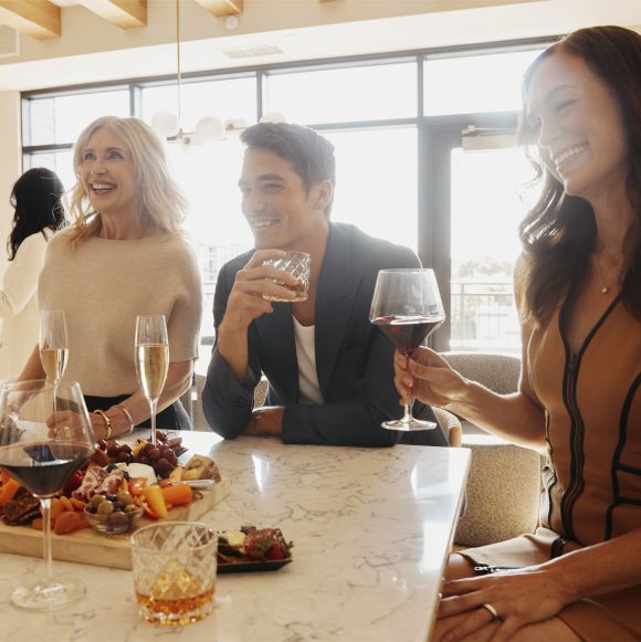 Three adults sit at a table, smiling and holding drinks, with a charcuterie board and glasses on the marble surface in front of them.