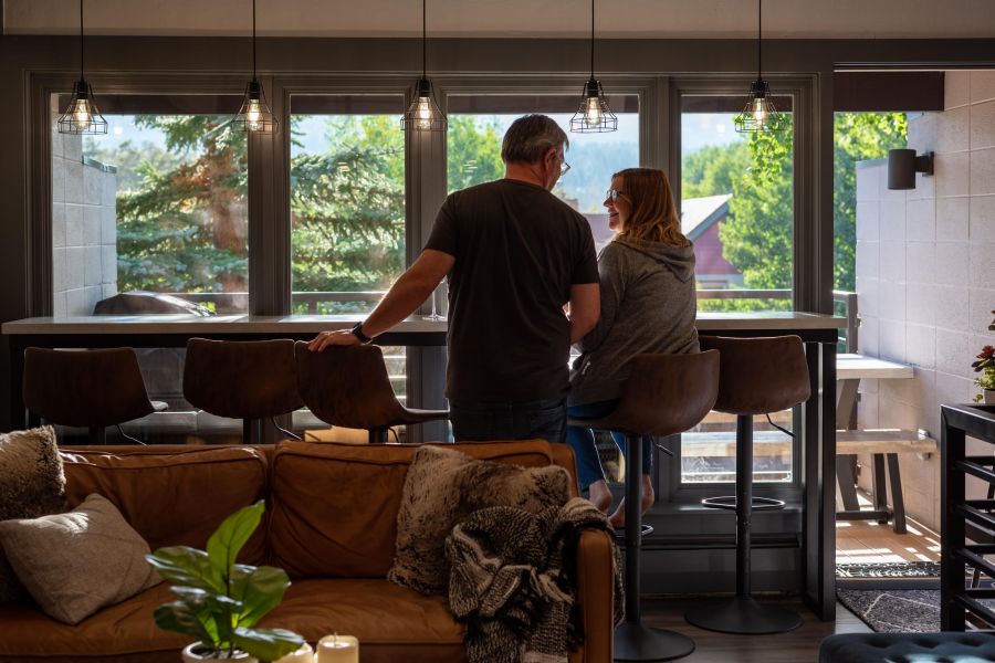 A man and woman talk at a kitchen island in a sunlit, modern living space with large windows and greenery outside.