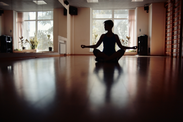 A person sits cross-legged meditating in a sunlit room with large windows and wooden floors.