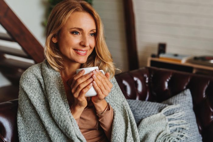 Smiling woman wrapped in a blanket sits on a couch holding a mug, looking cozy and relaxed.