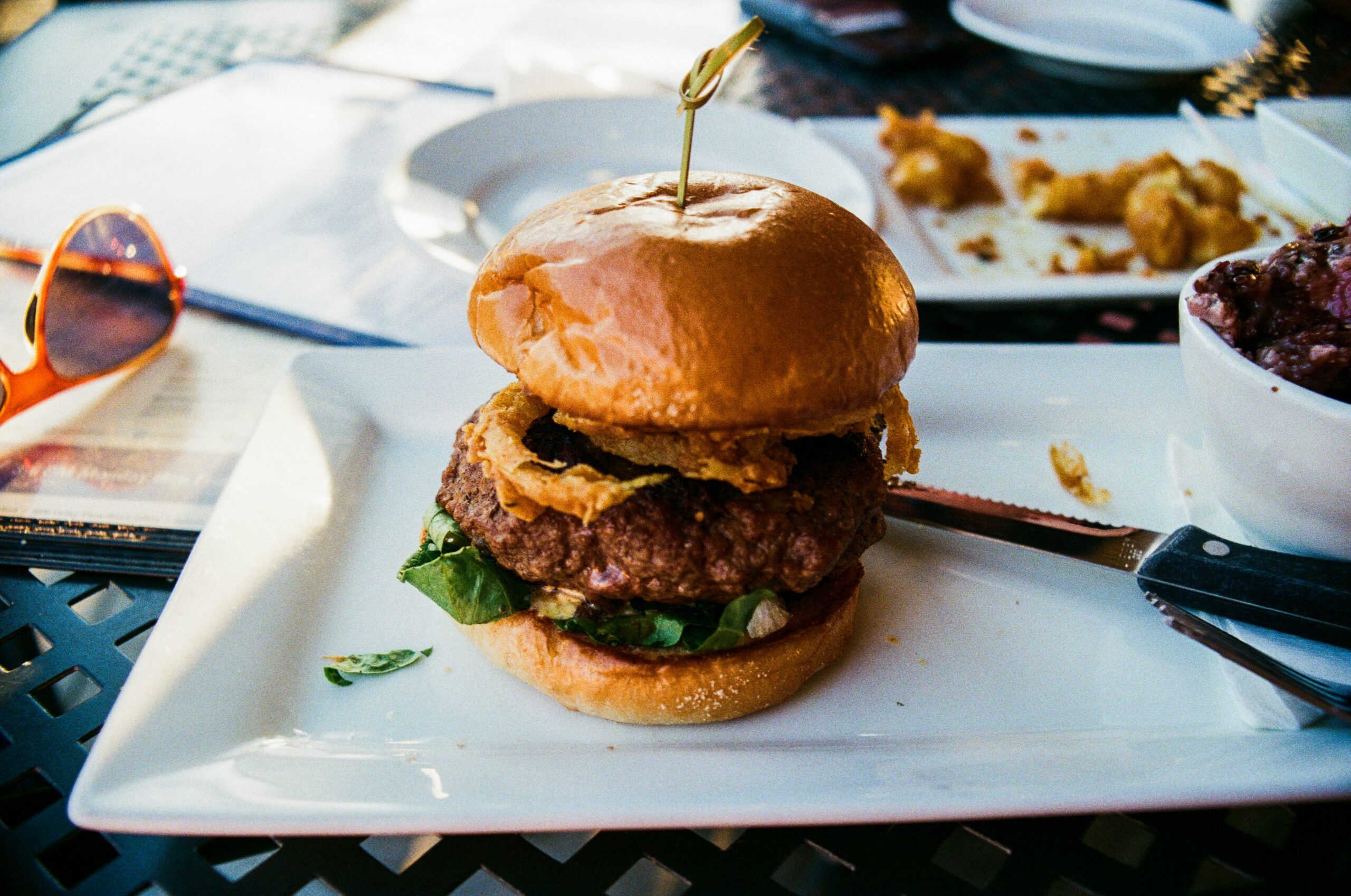 A burger with onion rings on a white plate, next to a knife and a ramekin, on an outdoor table.