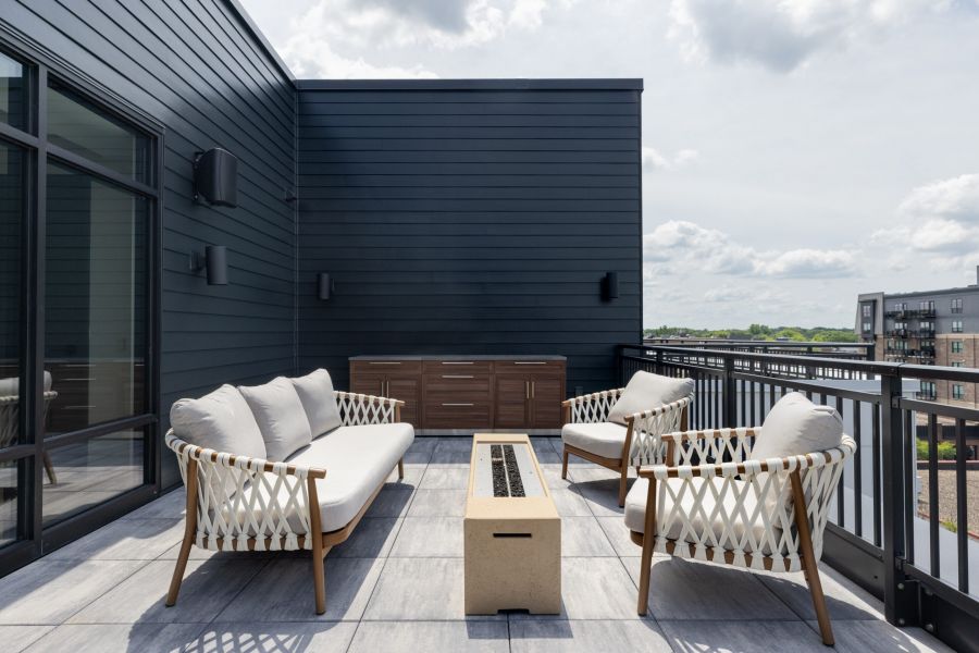 Modern rooftop patio with two chairs, a sofa, a fire table, outdoor cabinets, and a railing overlooking buildings.