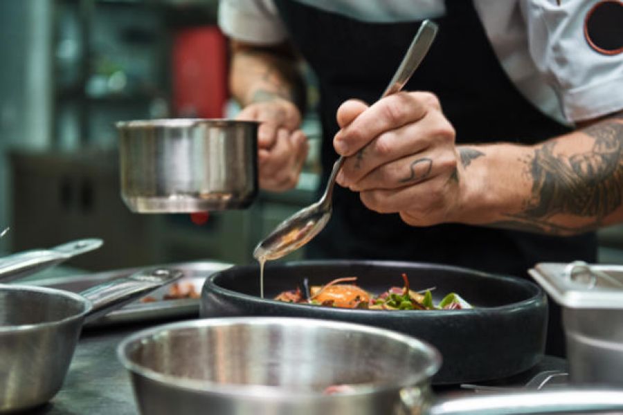 Chef plating a dish, pouring sauce with a spoon in a professional kitchen, metal pots in foreground.