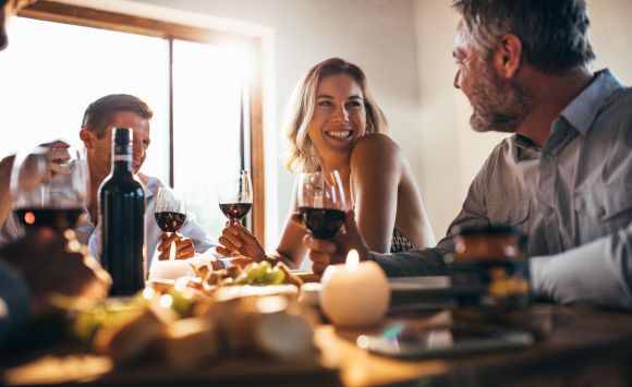 Four people sit at a table, smiling and drinking wine, with food and candles in the foreground.