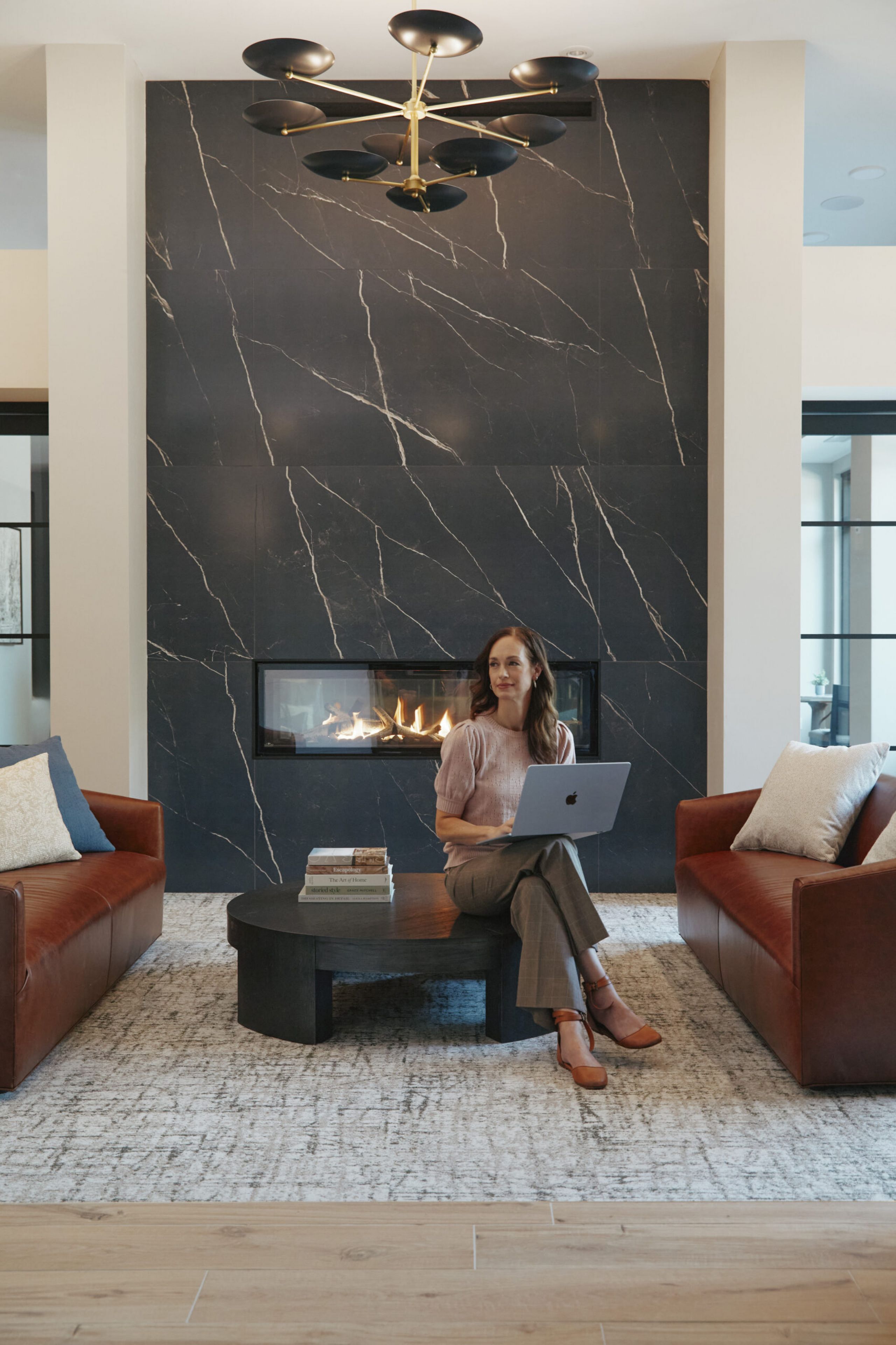 Woman sitting with a laptop on a coffee table between two brown sofas in a modern living room with a black marble fireplace.
