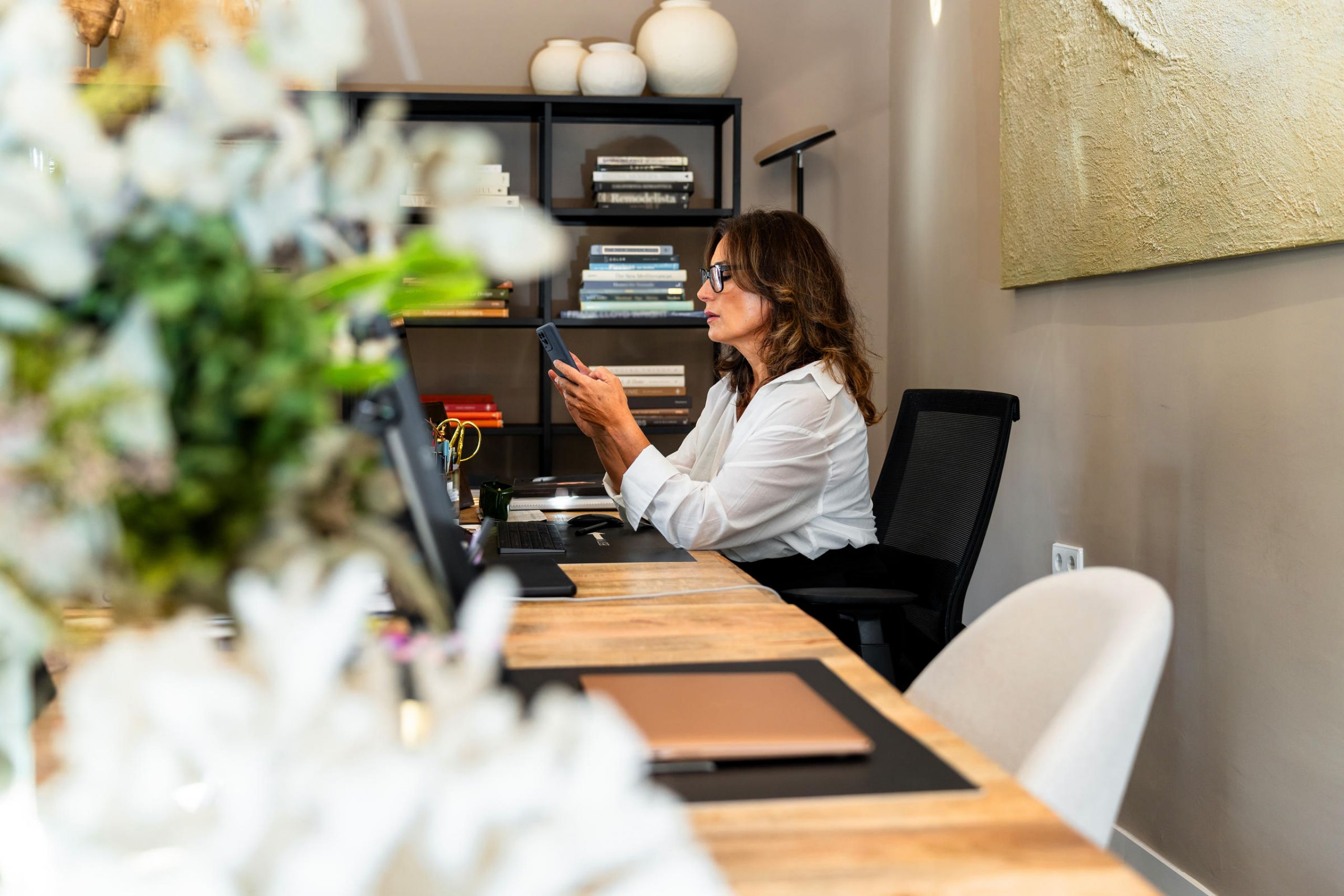 Woman in white shirt sitting at desk in modern office, looking at her phone, with shelves in background.