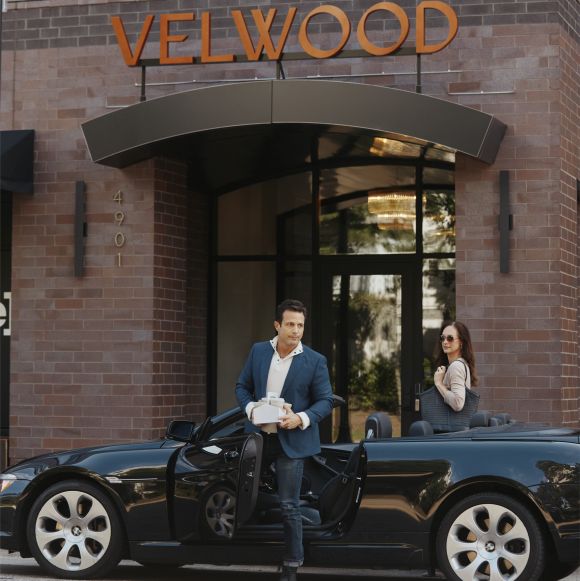 A man holds a box beside a black convertible while a woman sits inside, parked in front of a building labeled “VELWOOD.”.
