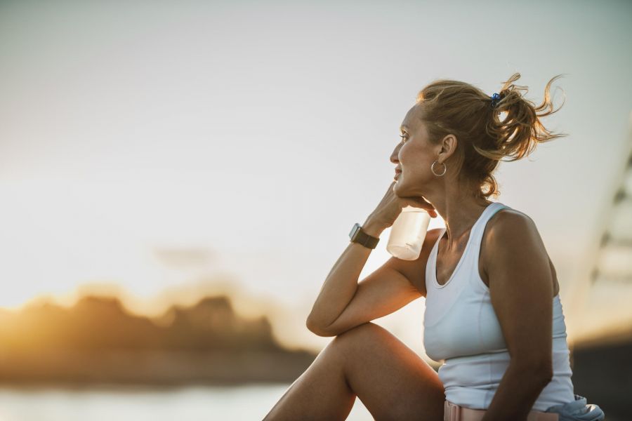 Woman in athletic wear sits outdoors at sunset, holding a water bottle and looking thoughtfully into the distance.