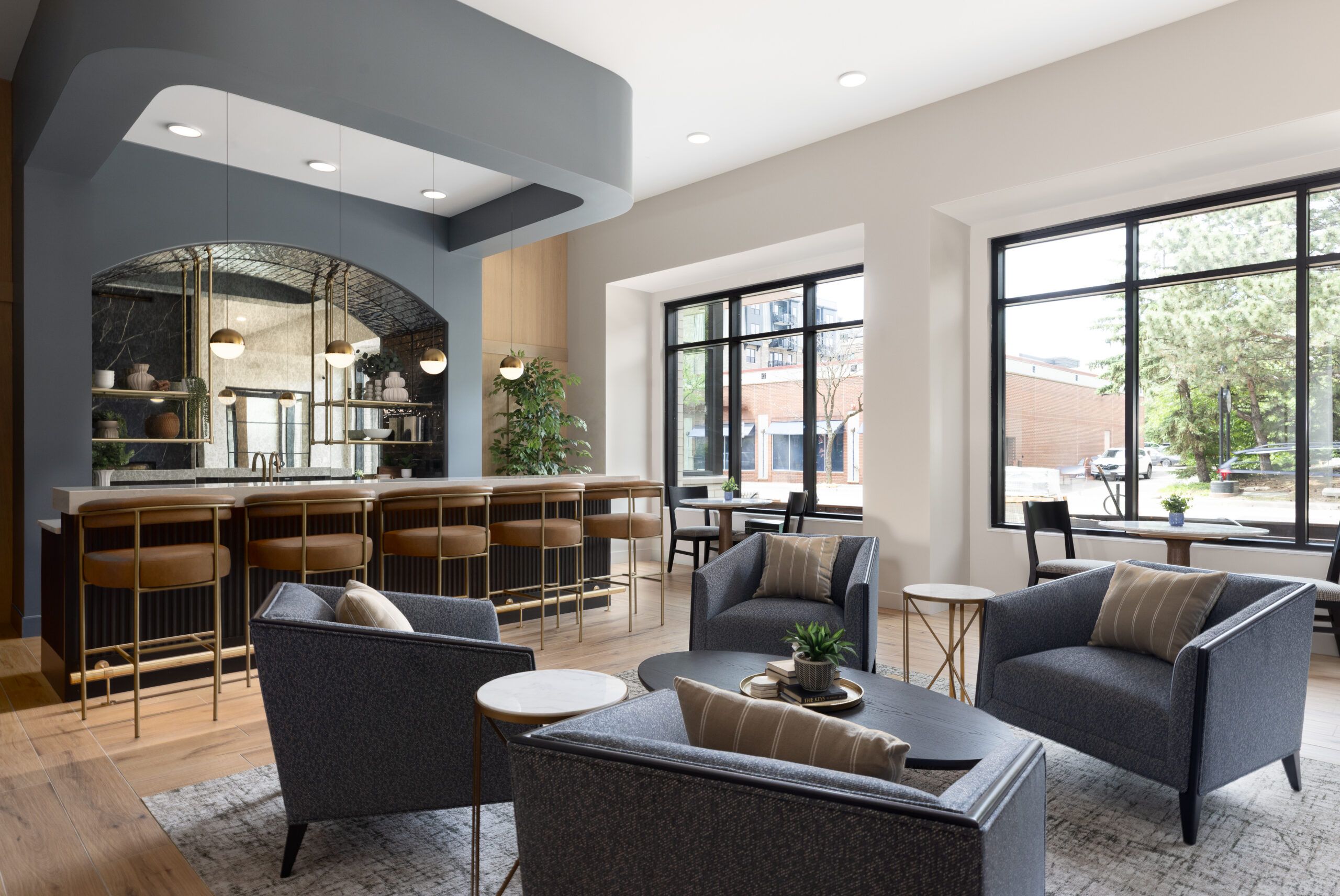 Modern lounge with gray armchairs, a bar area, large windows, and natural light filling the space.