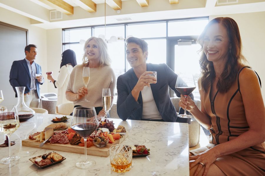 People enjoying drinks and appetizers around a kitchen island at a social gathering in a modern, sunlit home.