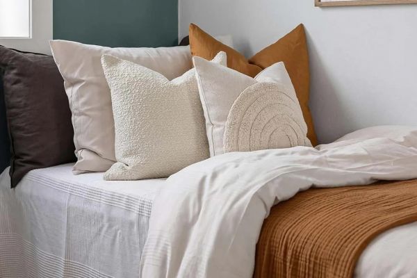 Cozy bed with beige, brown, and white pillows, white bedding, and a rust-colored blanket in a modern room.