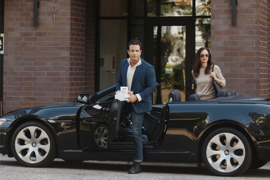 A man holding a drink exits a black convertible BMW as a woman sits in the passenger seat, in front of a brick building.