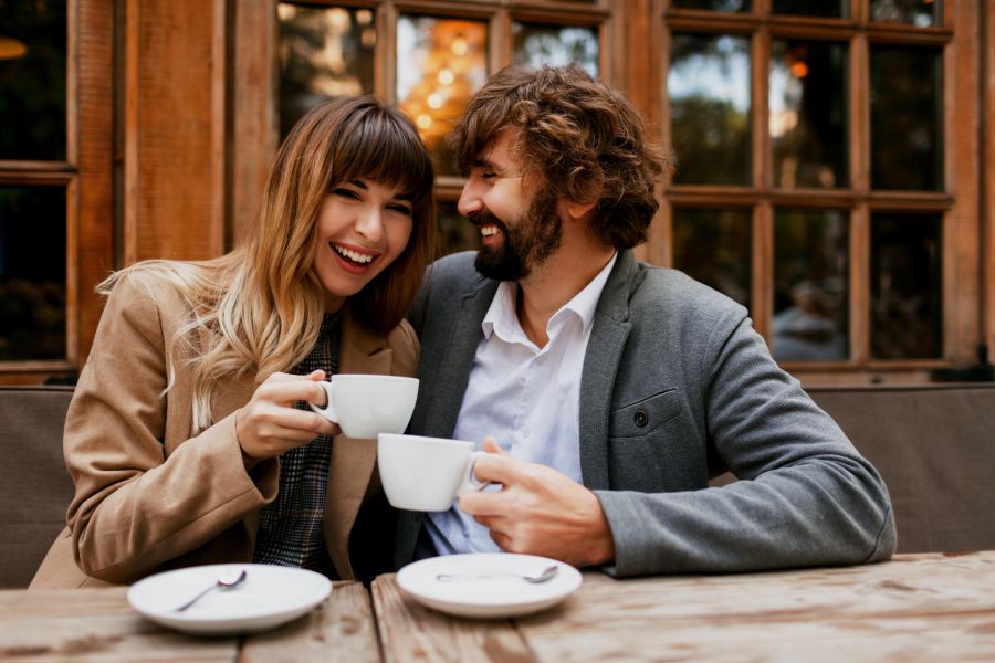 A smiling couple sits at an outdoor café, holding coffee cups and enjoying each other's company.
